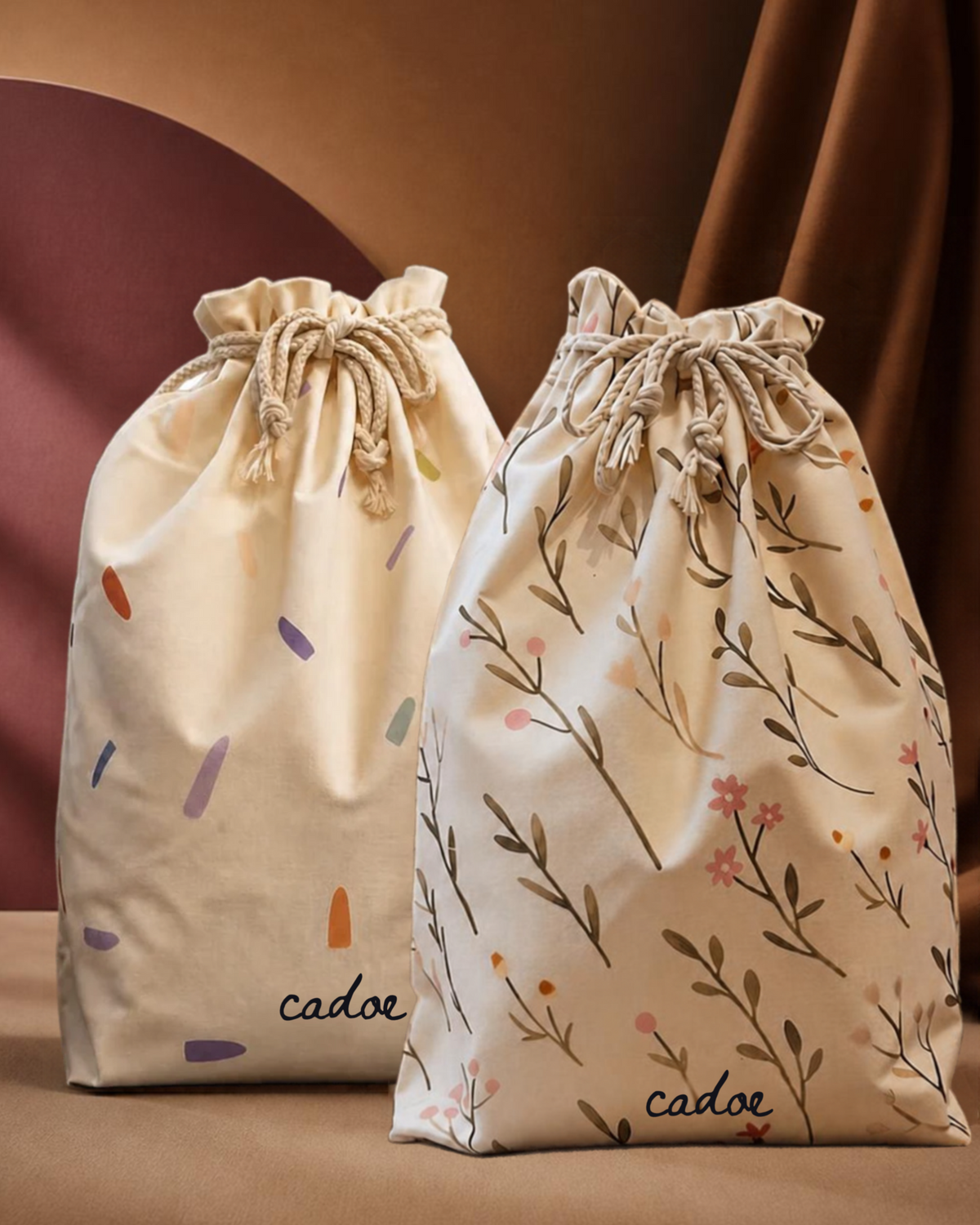 Two fabric drawstring pouches on a warm brown surface, one cream with scattered multicolor brushstrokes and the other cream with a floral branch print, both printed with the brand name 'cadoe'.